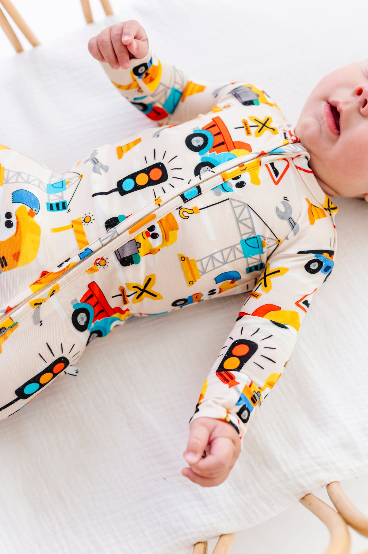 A baby lying on a white muslin mattress wearing a long-sleeve zip-up sleeper with a colorful construction vehicle print on a cream background.