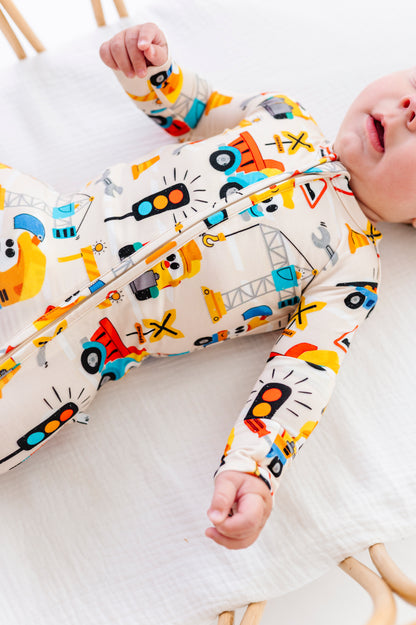 A baby lying on a white muslin mattress wearing a long-sleeve zip-up sleeper with a colorful construction vehicle print on a cream background.
