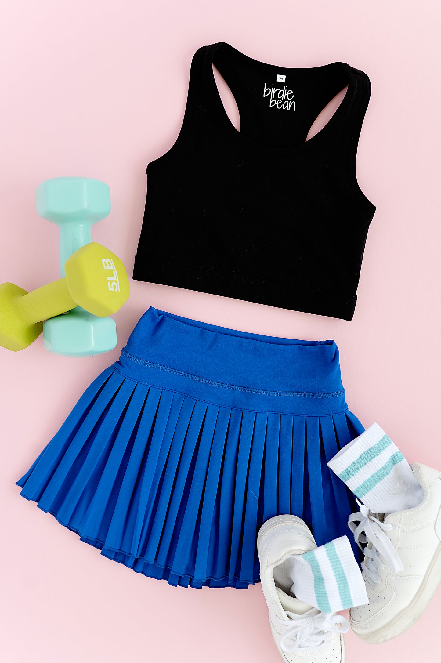 Black crop top, blue pleated skort, white sneakers, and green dumbbells on a pink background