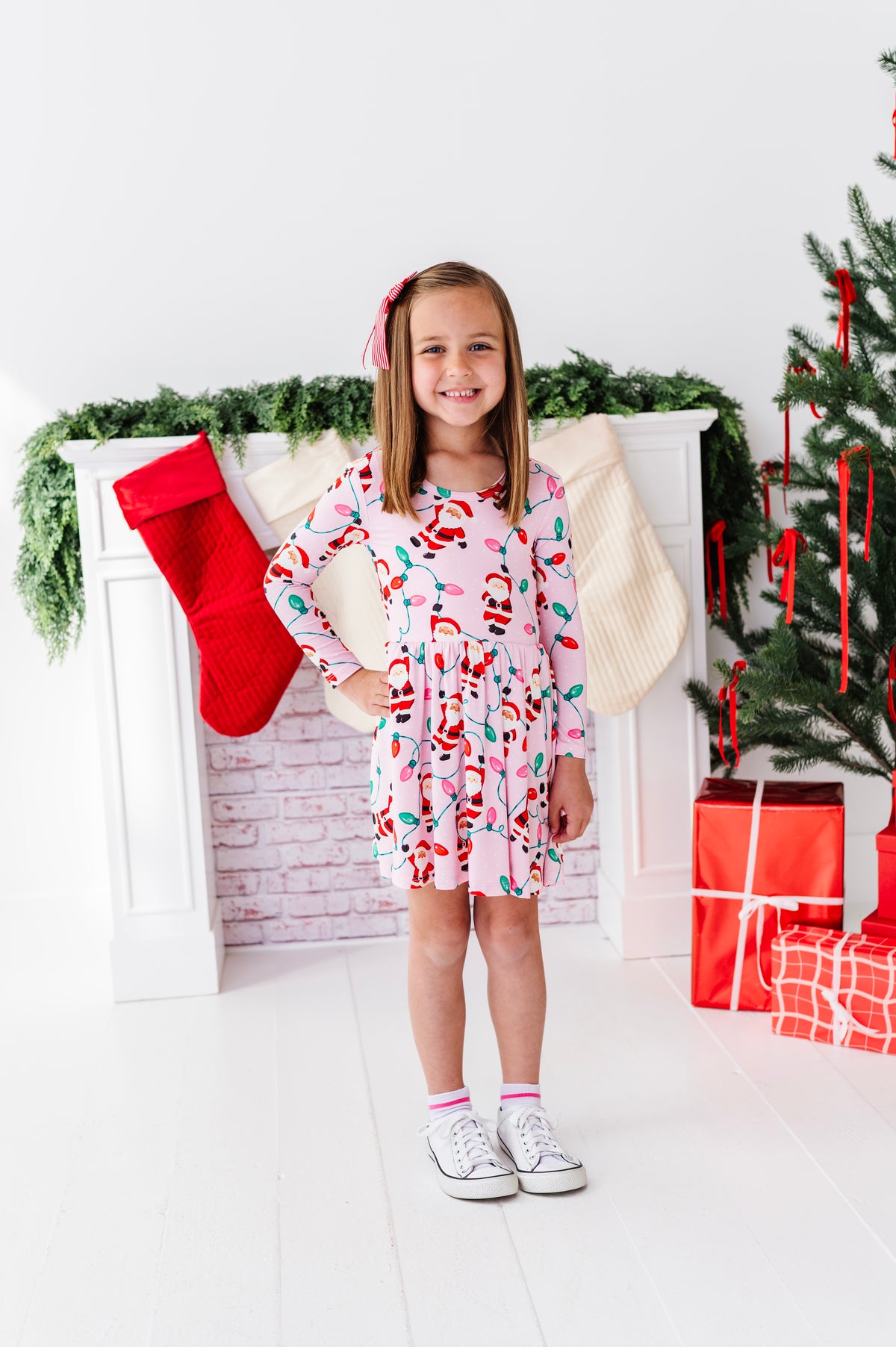 A young girl stands in a holiday-themed room wearing a pink long-sleeve bamboo dress with a colorful Santa Claus pattern. The background features a white fireplace mantel with a red stocking, green garland, and a Christmas tree with wrapped presents.