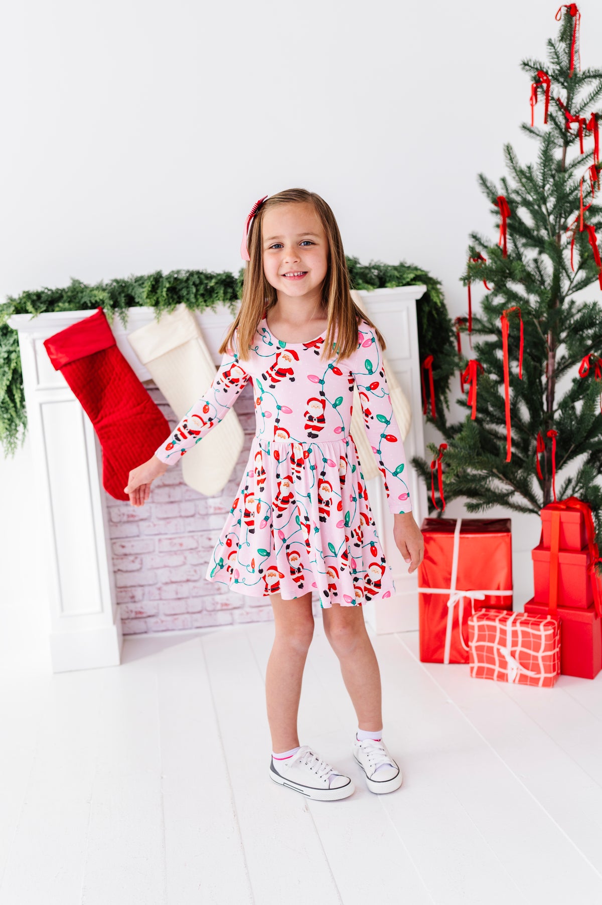 A young girl stands in a holiday-themed room wearing a pink long-sleeve bamboo dress with a Santa Claus print. The background features a white fireplace with stockings, a Christmas tree, and wrapped gifts.