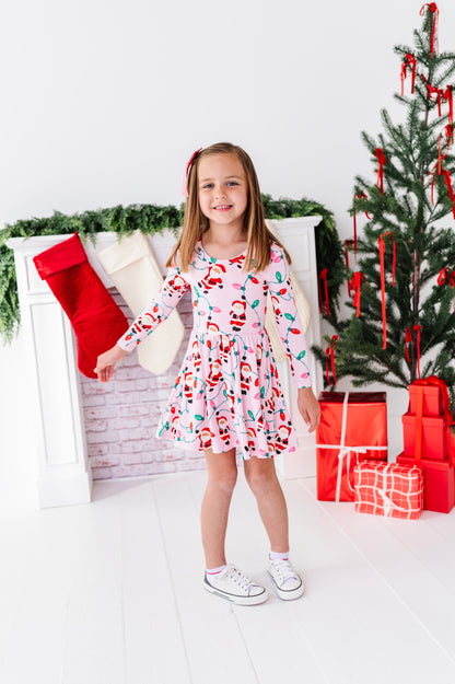 A young girl stands in a holiday-themed room wearing a pink long-sleeve bamboo dress with a Santa Claus print. The background features a white fireplace with stockings, a Christmas tree, and wrapped gifts.