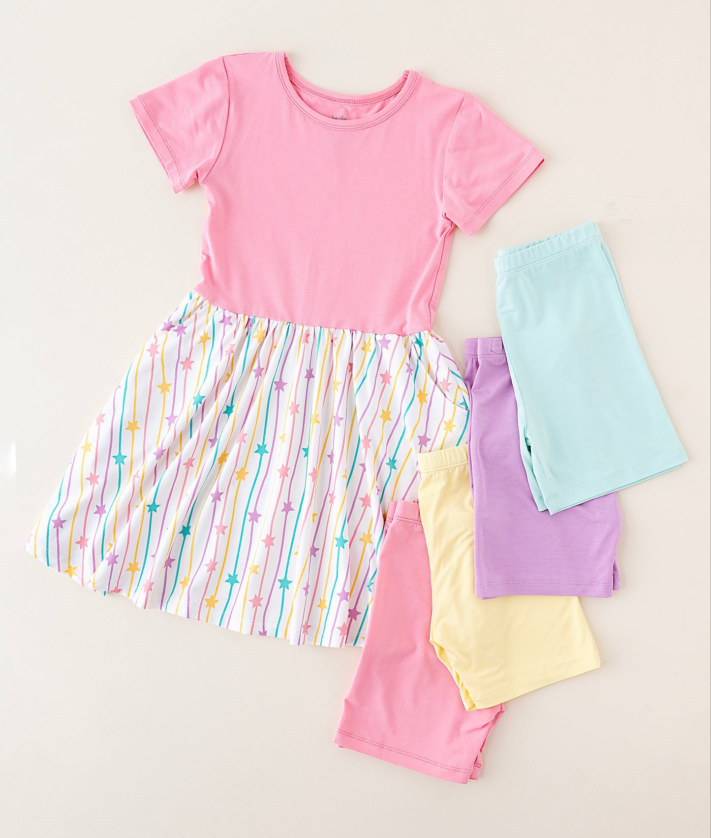 A children's short-sleeve dress laid flat on a light beige background, featuring a solid pink upper bodice and a gathered multicolor striped skirt with pastel rainbow lines and small star motifs. Four folded matching shorts or pant legs in light blue, lavender, pale yellow, and pink are arranged beside the dress.