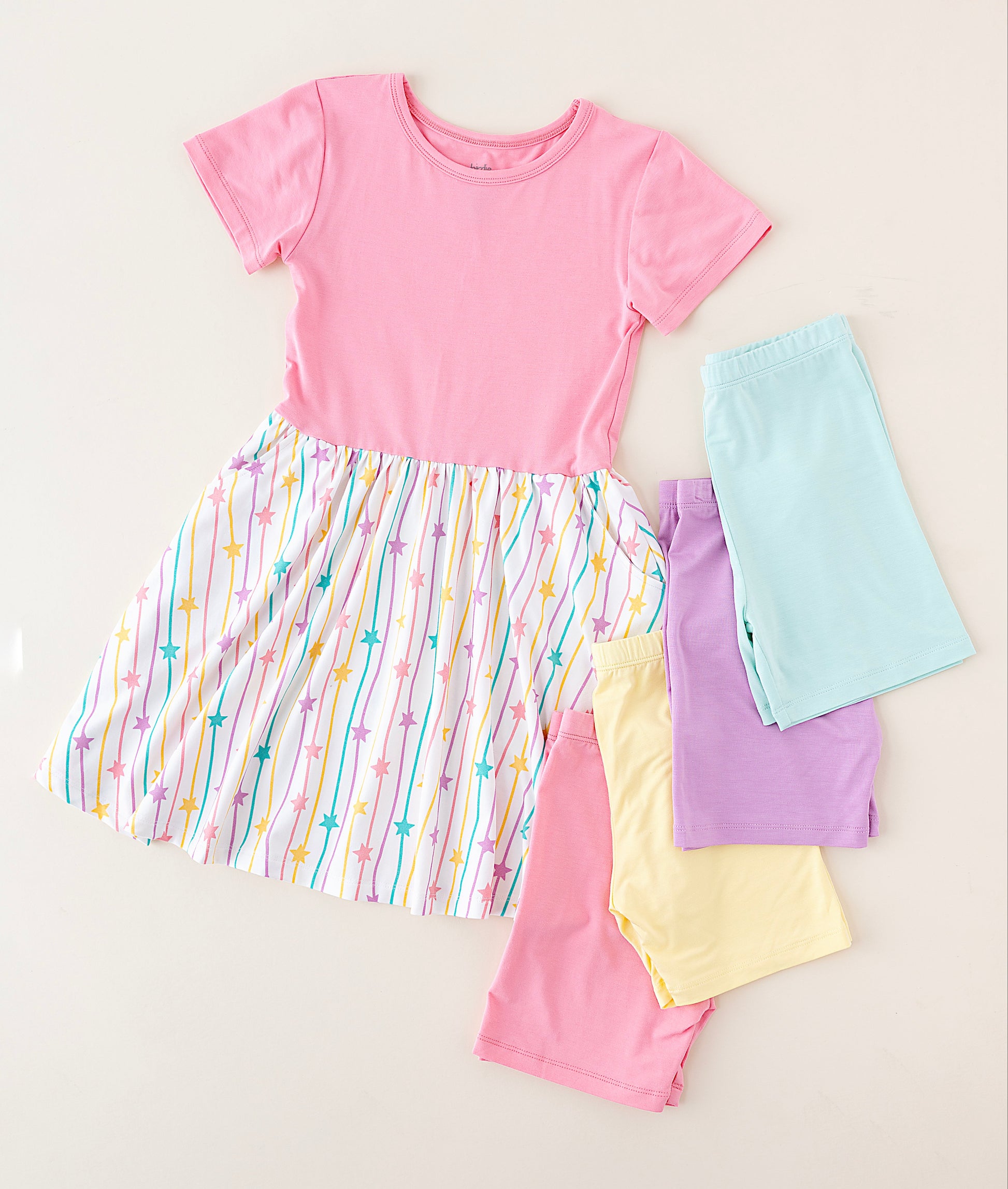 A children's short-sleeve dress laid flat on a light beige background, featuring a solid pink upper bodice and a gathered multicolor striped skirt with pastel rainbow lines and small star motifs. Four folded matching shorts or pant legs in light blue, lavender, pale yellow, and pink are arranged beside the dress.