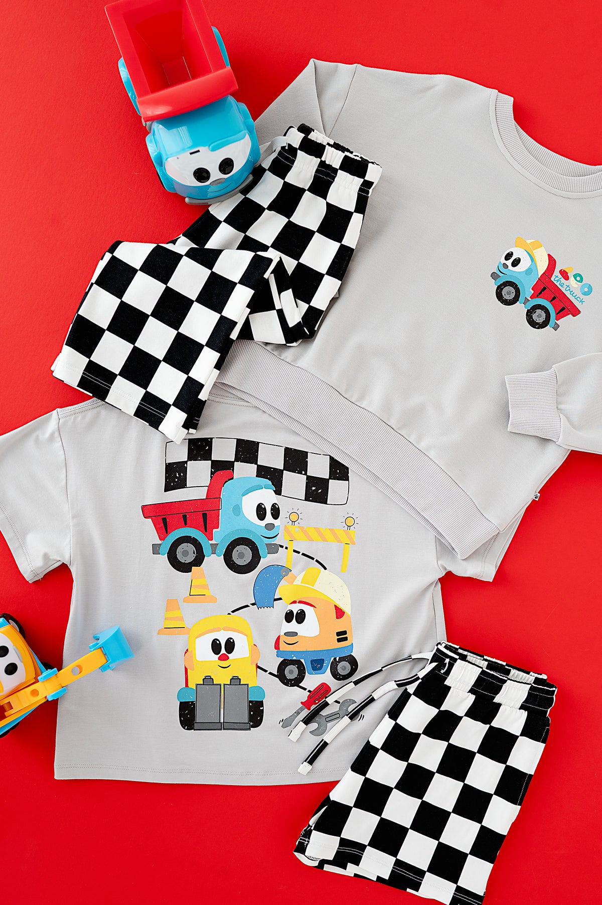 A children's two-piece pajama set laid flat on a bright red background, featuring a light gray long-sleeve top and matching shorts with black-and-white checkered patterns. The tops are printed with colorful cartoon construction vehicles, and the shorts have a checkered border.