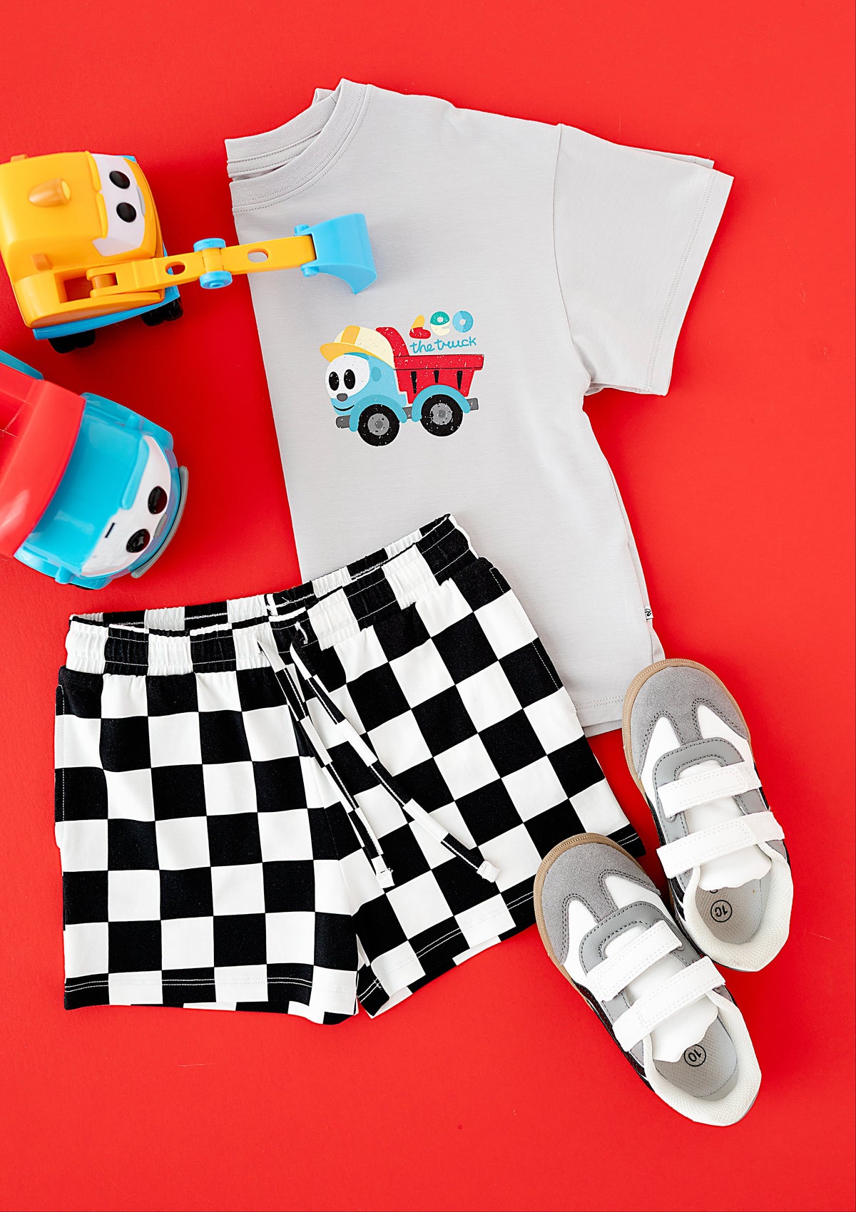A children's outfit flat lay on a bright red background, featuring a white short-sleeve T-shirt with a cartoon dump truck graphic, black-and-white checkered shorts, gray and white velcro sneakers, and toy vehicles.