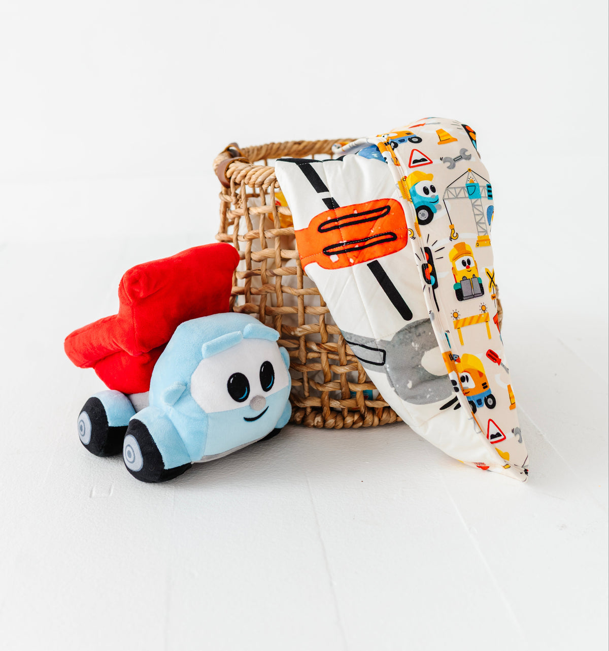 A woven wicker basket with a triangular fabric cover printed with colorful construction vehicles, shown next to a plush toy car on a clean white background.