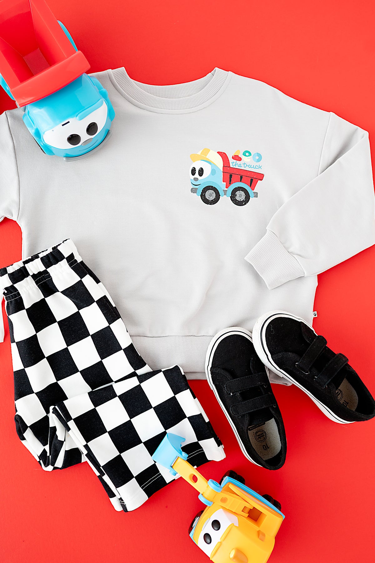 A children's outfit flat lay on a bright red background, featuring a white sweatshirt with a colorful dump truck graphic, black-and-white checkered pants, black velcro sneakers, and toy vehicles.