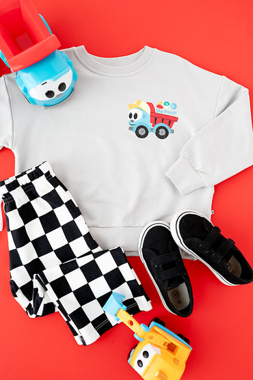 A children's outfit flat lay on a bright red background, featuring a white sweatshirt with a colorful dump truck graphic, black-and-white checkered pants, black velcro sneakers, and toy vehicles.