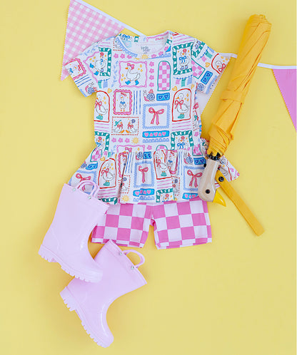 Children's outfit with a patterned top and checkered shorts, accompanied by an umbrella and rain boots on a yellow background.