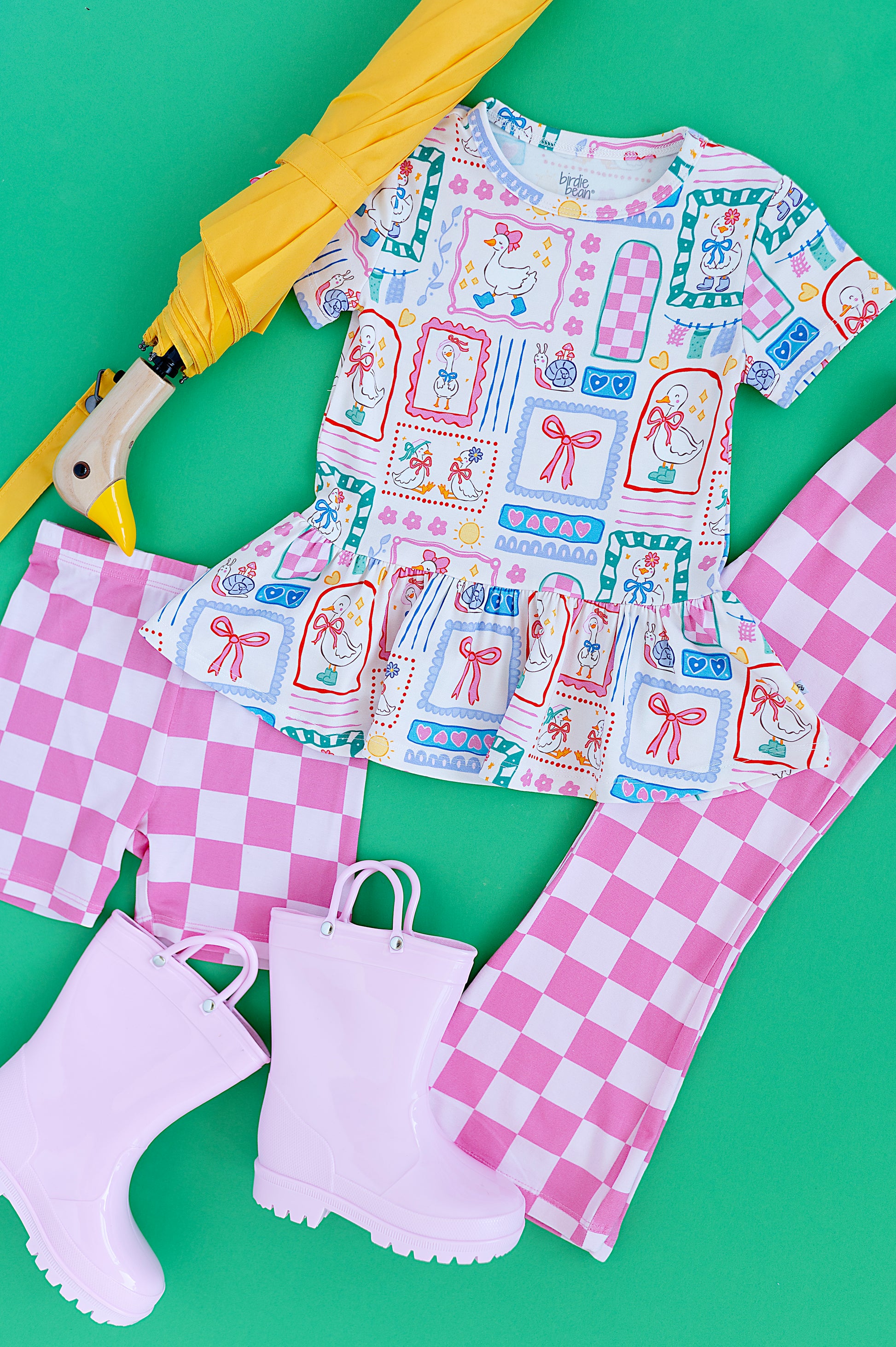 Children's outfit with patterned Gilly Goose Bamboo/Cotton Peplum Tee, bamboo checkered shorts, bamboo checkered flare pants, rain boots, and an umbrella on a green background