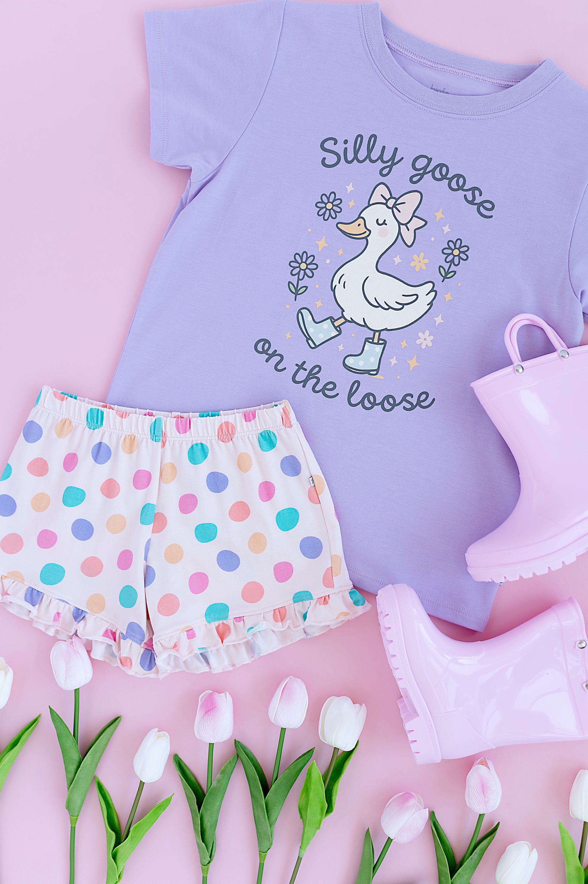 Children's outfit with a purple shirt, polka dot shorts, and pink boots on a pink background with tulips.