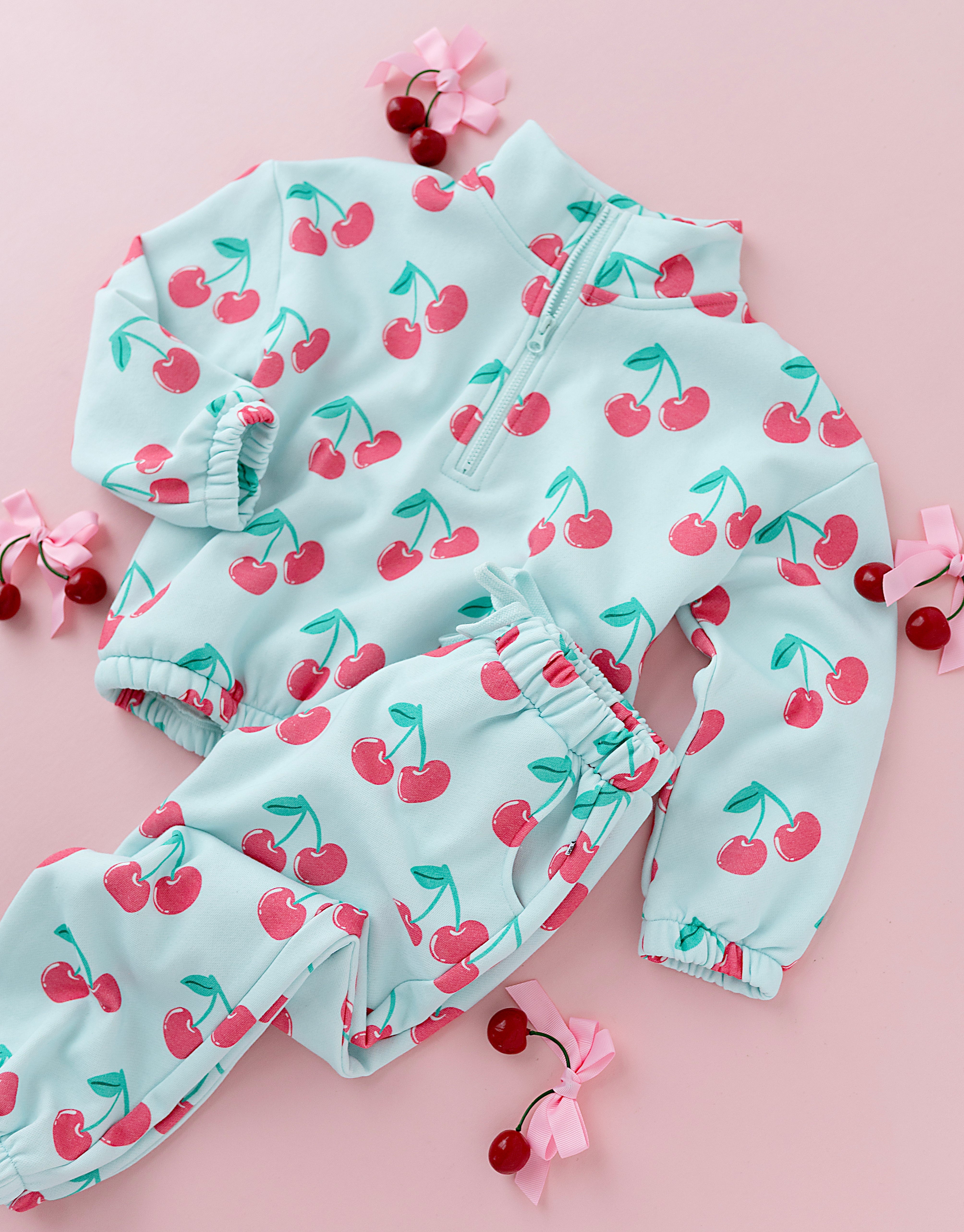 A baby two-piece outfit set consisting of a light blue zip-up jacket and matching pants, both covered in a red cherry print, arranged on a pink background with decorative cherry props.