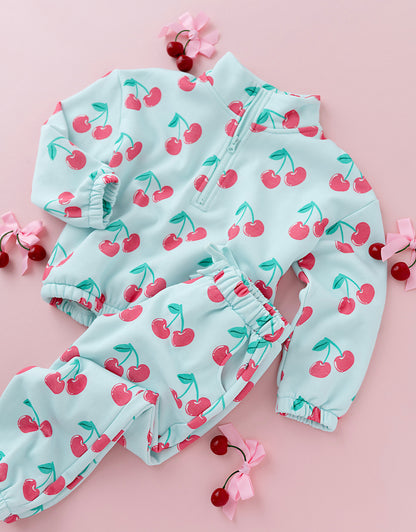A baby two-piece outfit set consisting of a light blue zip-up jacket and matching pants, both covered in a red cherry print, arranged on a pink background with decorative cherry props.