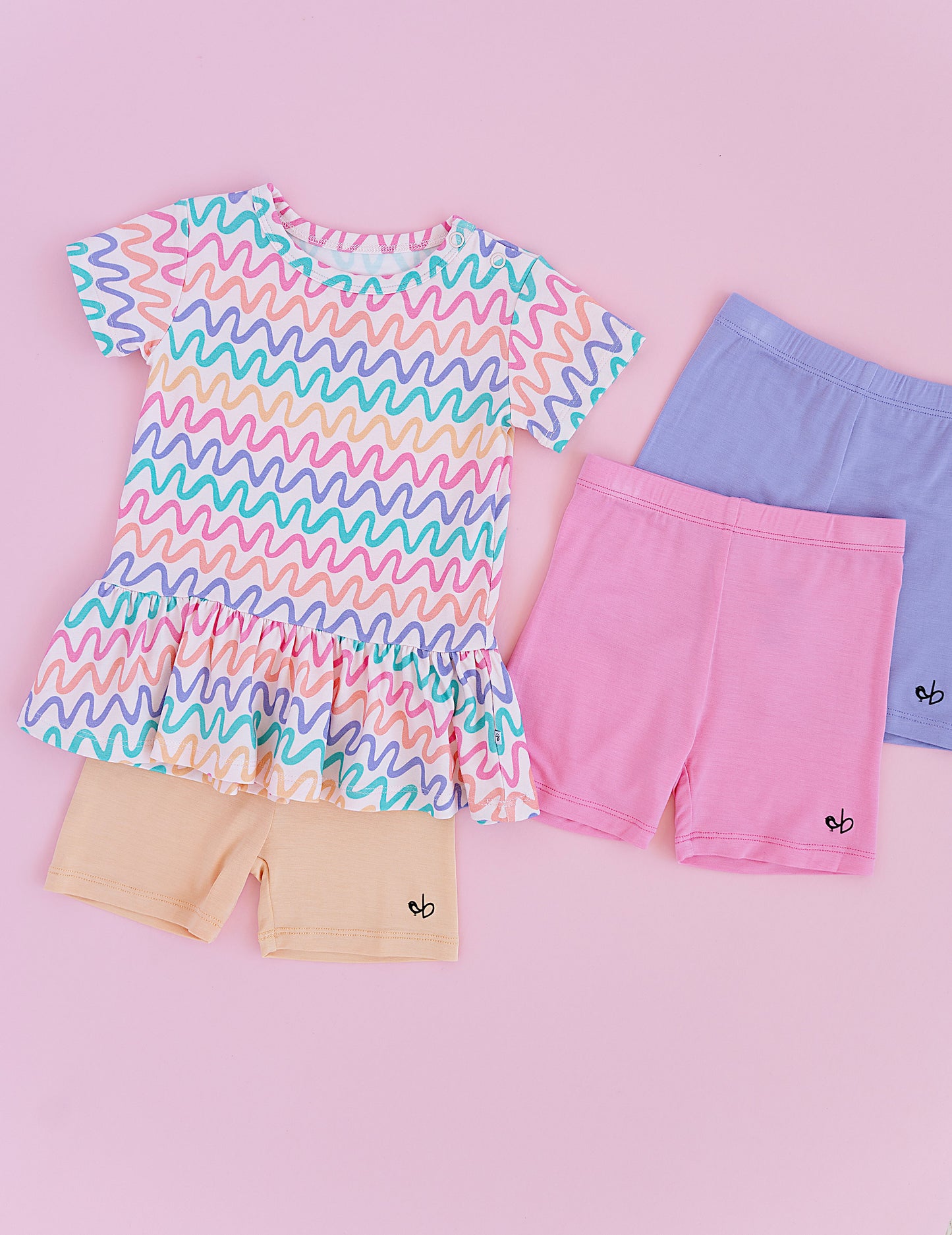 Children's clothing with a colorful wavy top, and coordinating biker shorts in the colors pink, purple, and yellow on a pink background.