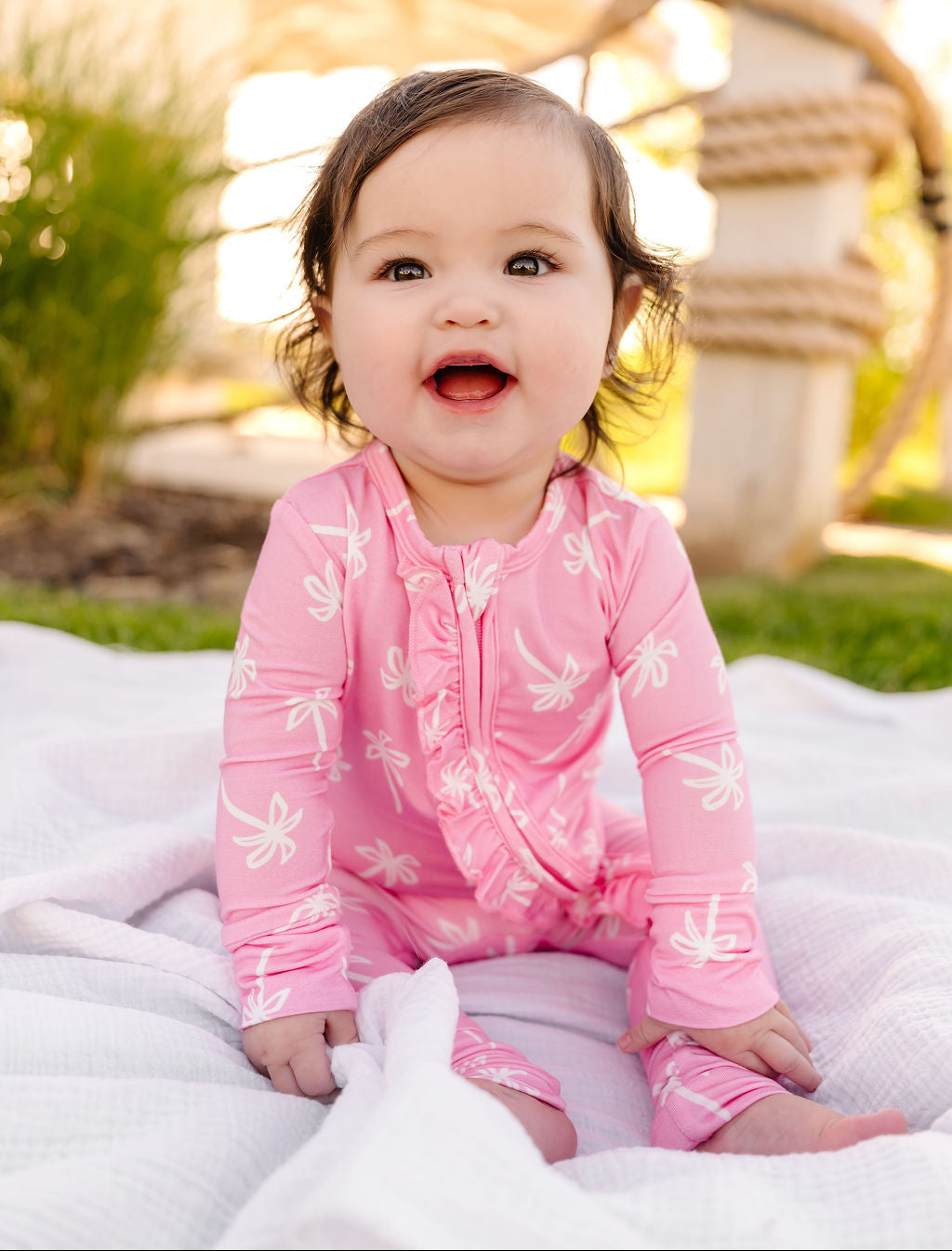 Baby in a pink palm outfit sitting on a blanket outdoors.