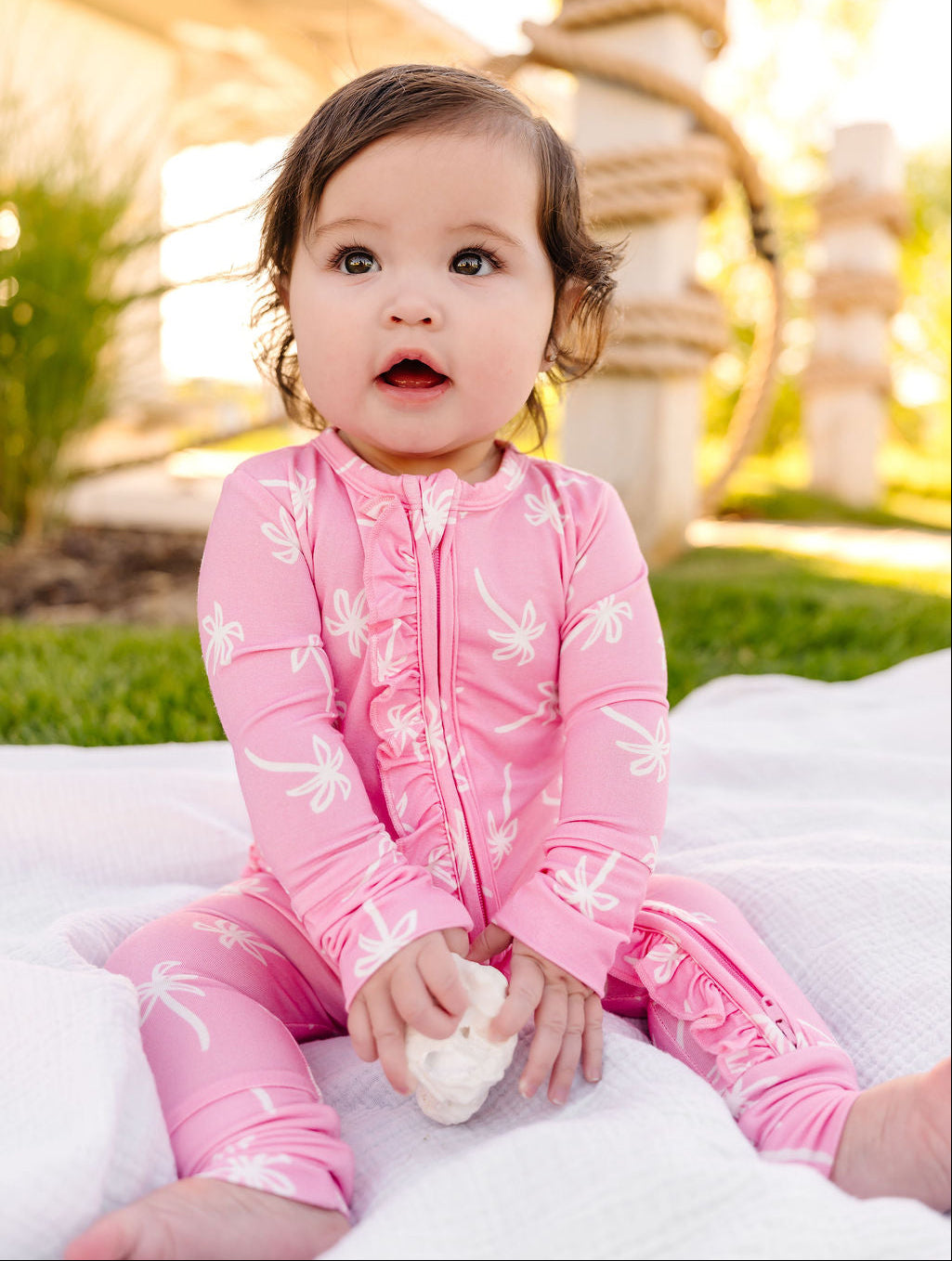 Child wearing a pink outfit with white patterns, sitting on a white blanket outdoors.
