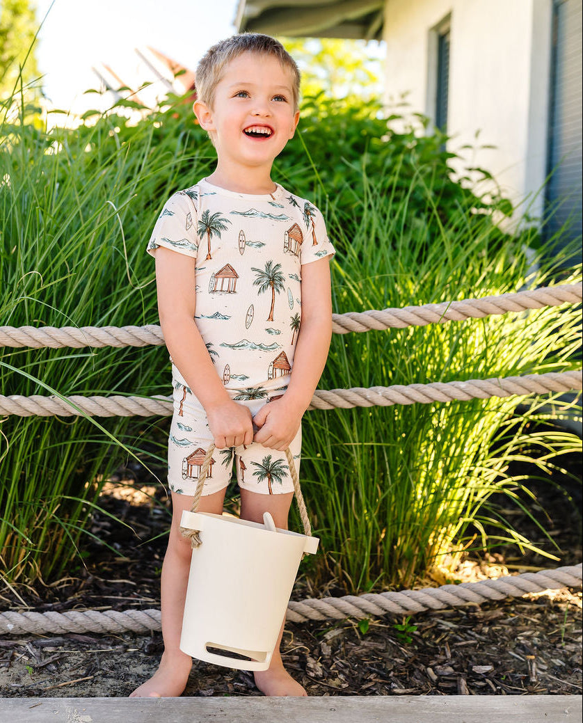 Child in a palm tree-themed outfit holding a bucket on a wooden deck with greenery in the background