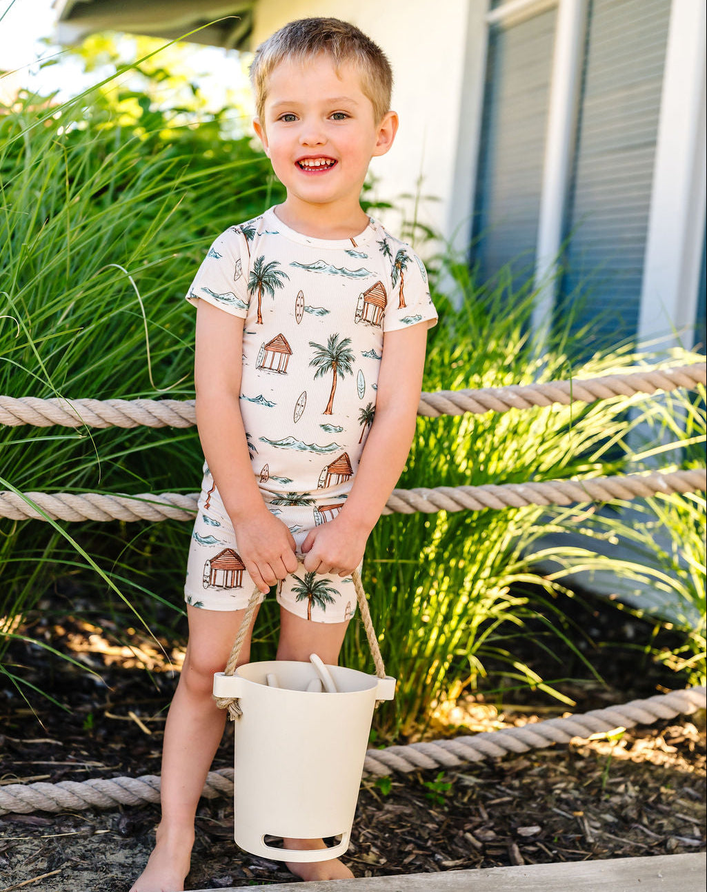 Young boy in a tropical-themed pj set holding a sand bucket outdoors.