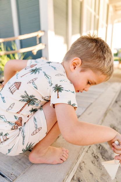 Child in a palm tree patterned outfit sitting on a wooden surface.