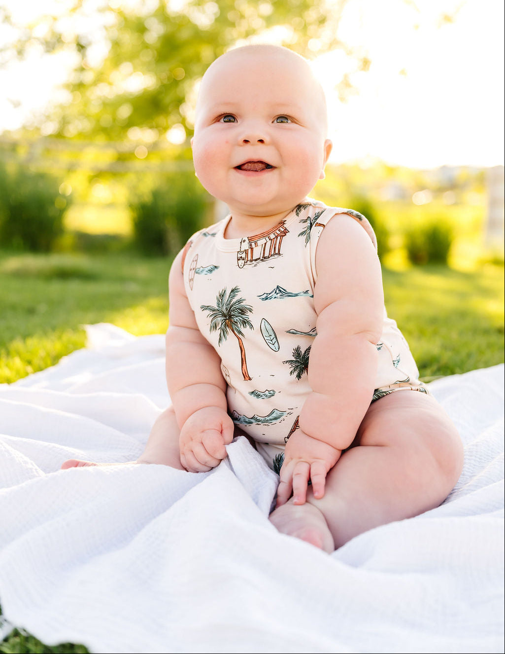 Baby sitting on a white blanket in a park with trees and grass in the background