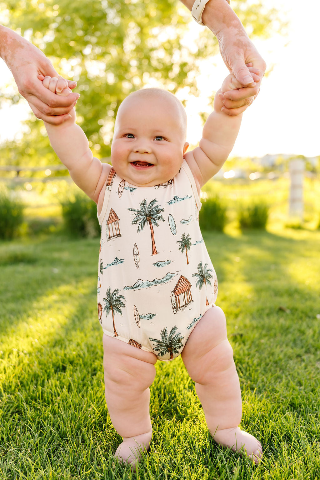 Baby in a onesie with palm tree design standing on grass, holding hands with adults.