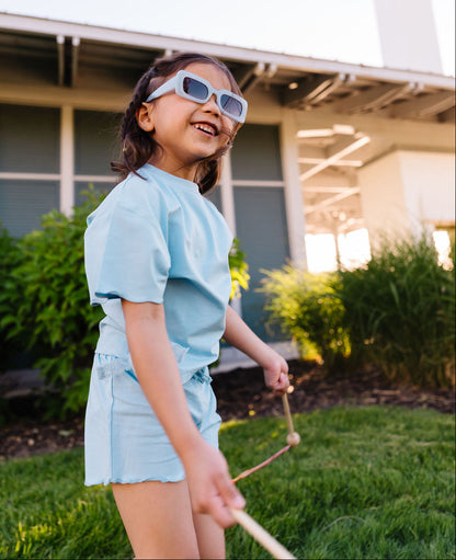 Child wearing sunglasses and a light blue outfit standing outdoors.