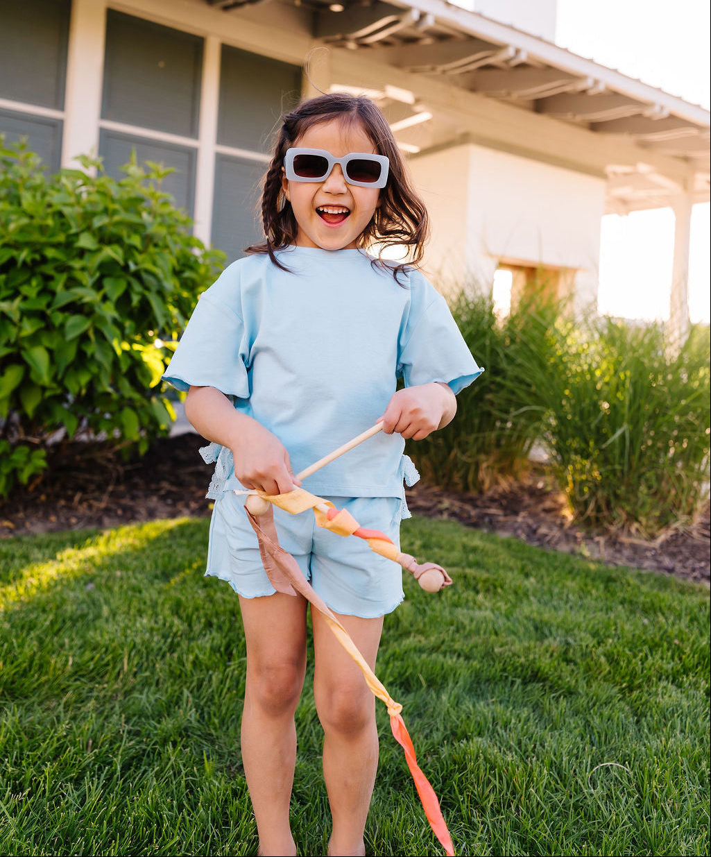Child wearing sunglasses and a light blue outfit standing on grass with a house in the background