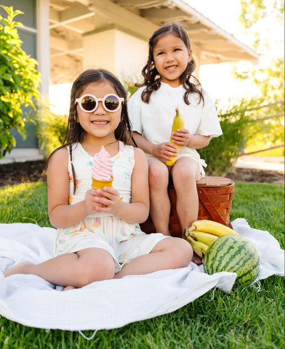 Two young girls enjoying ice cream and bananas on a picnic blanket in a park.