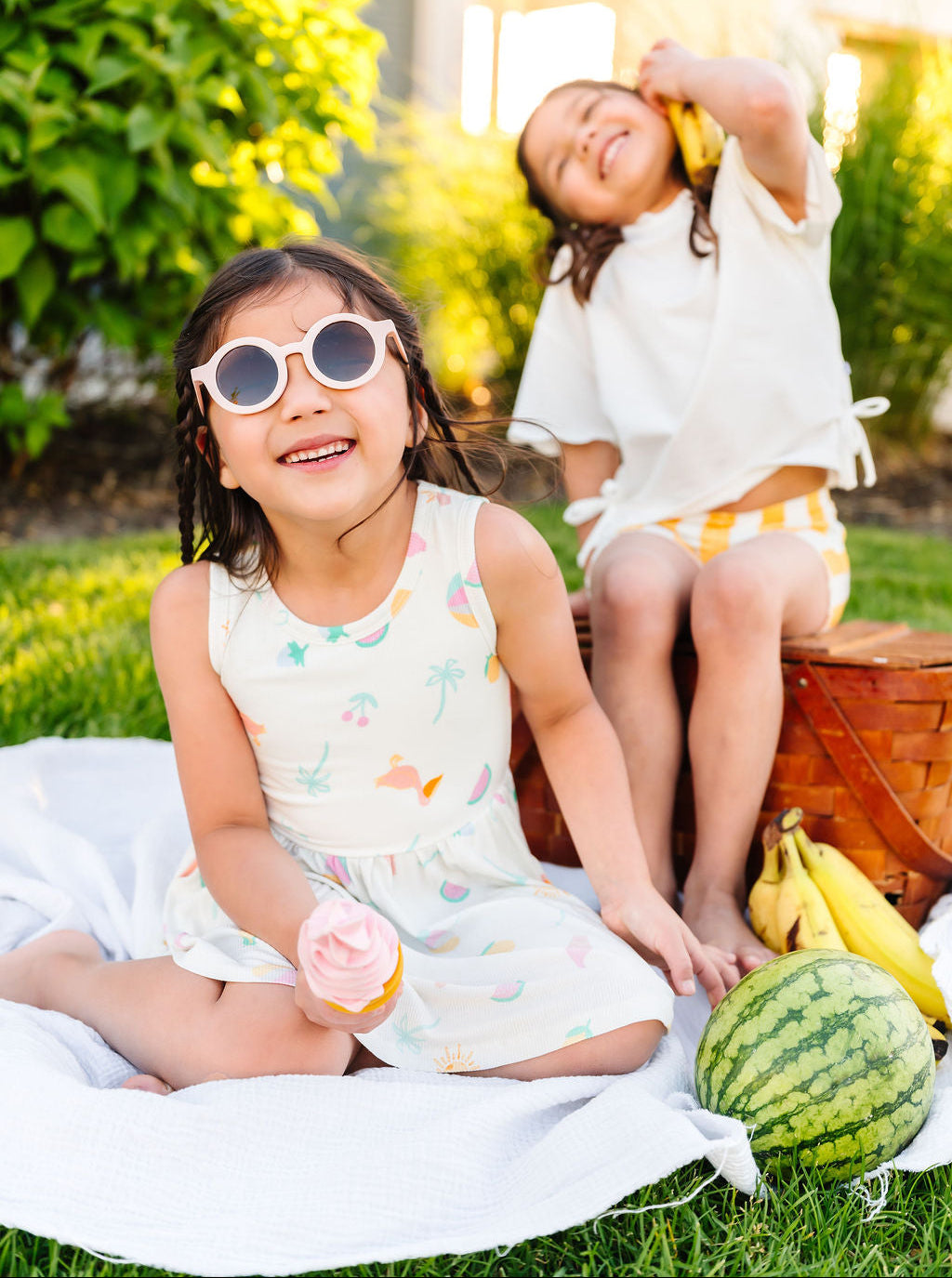 Two children sitting on a blanket outdoors with a watermelon and bananas.