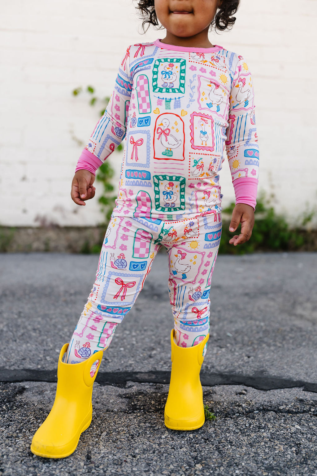 Child wearing a colorful patterned outfit with yellow rain boots on a concrete surface.
