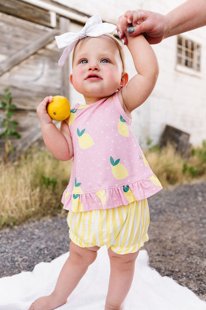 Baby in a pink outfit with lemon design holding a lemon outdoors.