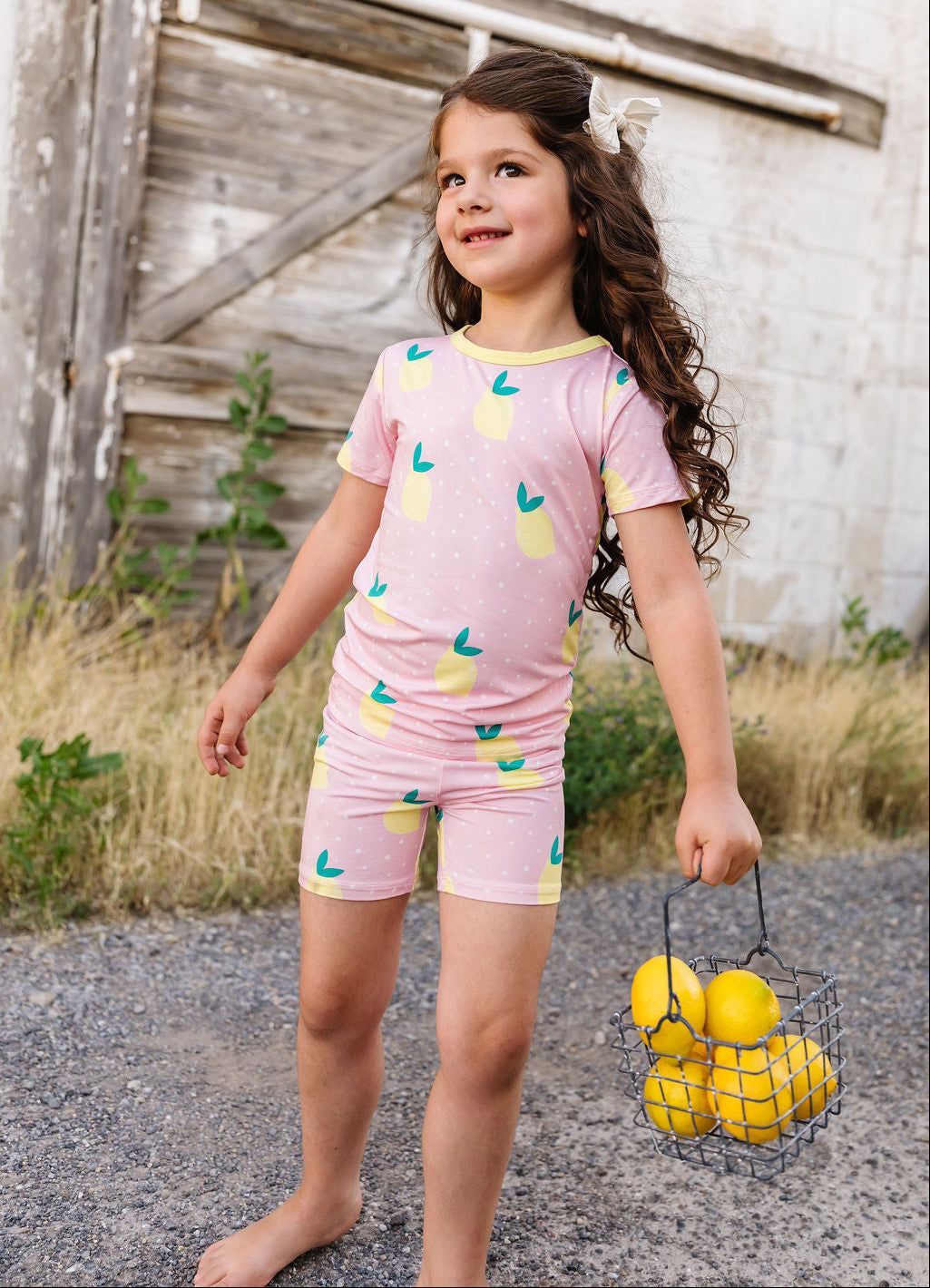 Young girl in a pink outfit with lemon pattern holding a basket of lemons outdoors.