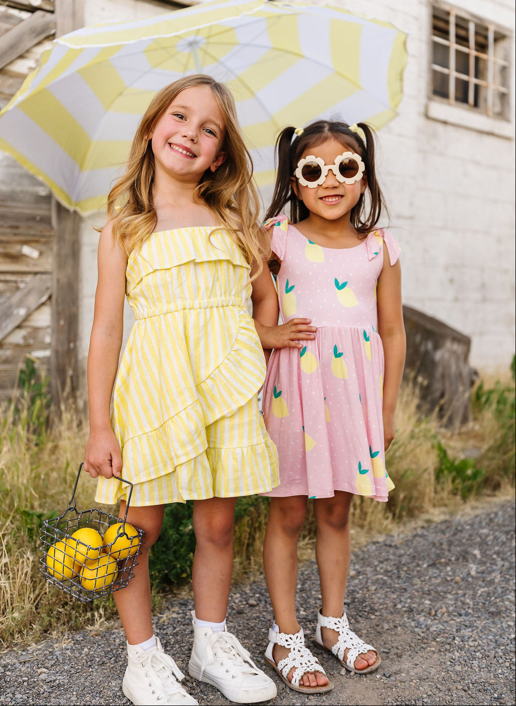 Two young girls in colorful dresses standing outdoors with a yellow and white umbrella.