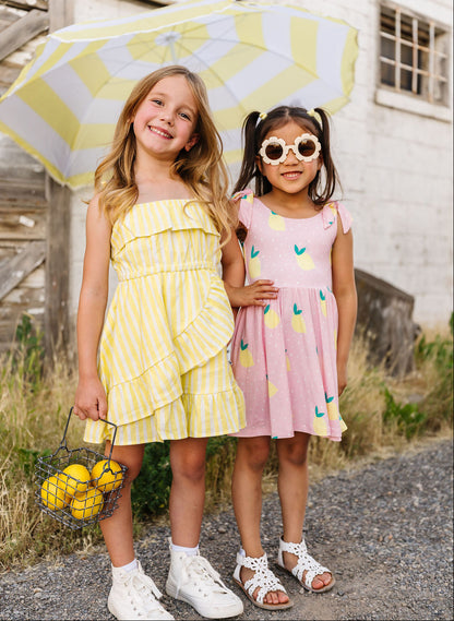Two young girls in colorful dresses standing outdoors with a yellow and white umbrella.
