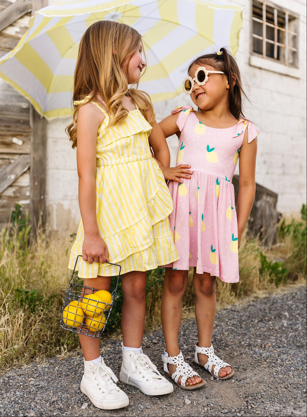 Two young girls standing under a yellow and white striped umbrella, wearing dresses and white shoes.