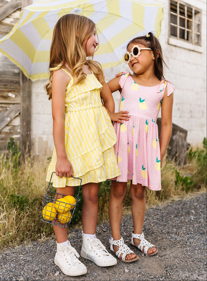 Two young girls standing under a yellow and white striped umbrella, wearing dresses and white shoes.