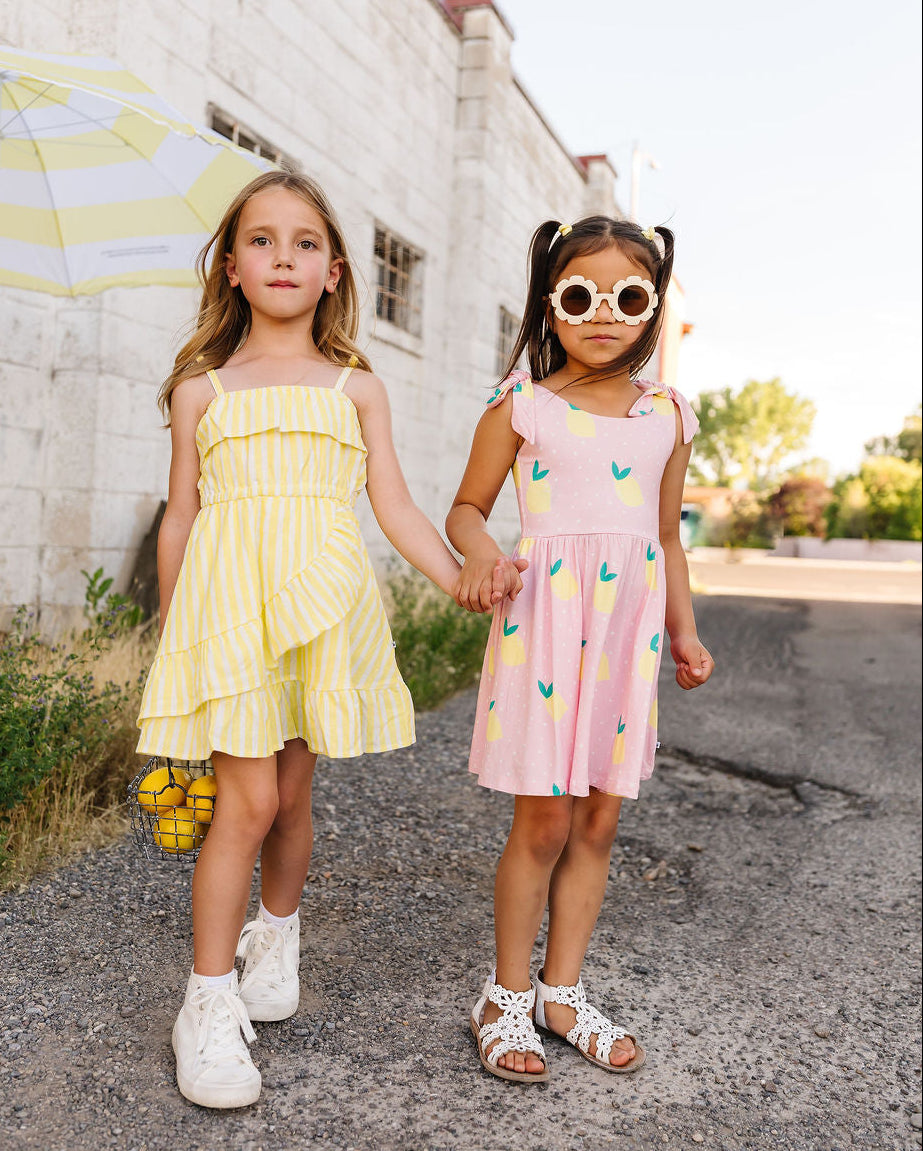 Two young girls holding hands, one in a yellow dress and the other in a pink dress with fruit pattern, standing on a street.