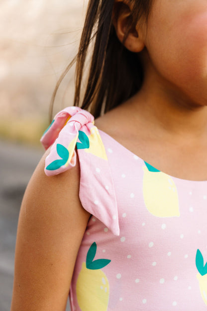 Child wearing a pink dress with yellow and lemonpatterns, standing outdoors.
