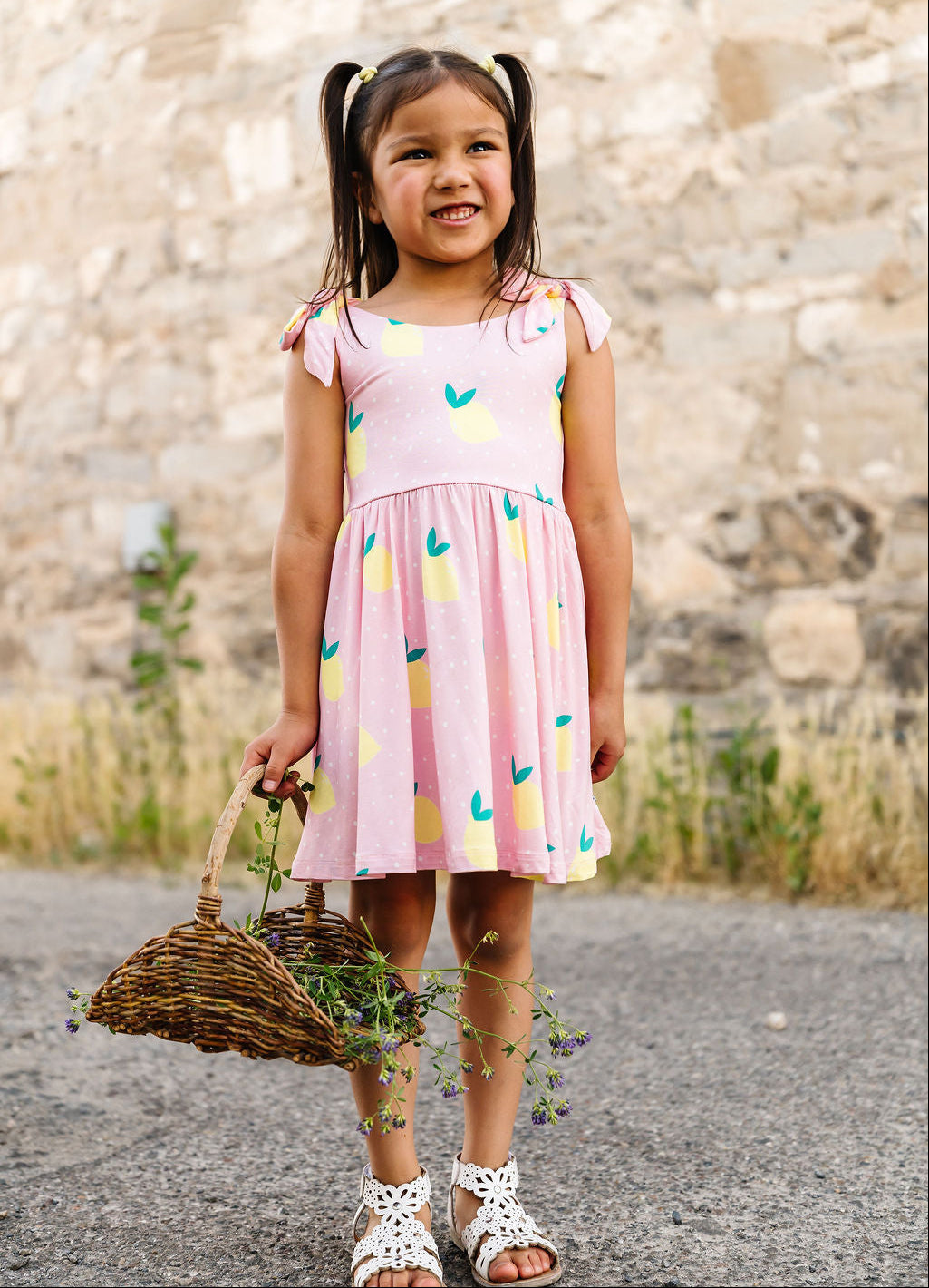 Young girl in a pink dress with colorful patterns, holding a basket, standing against a stone wall.