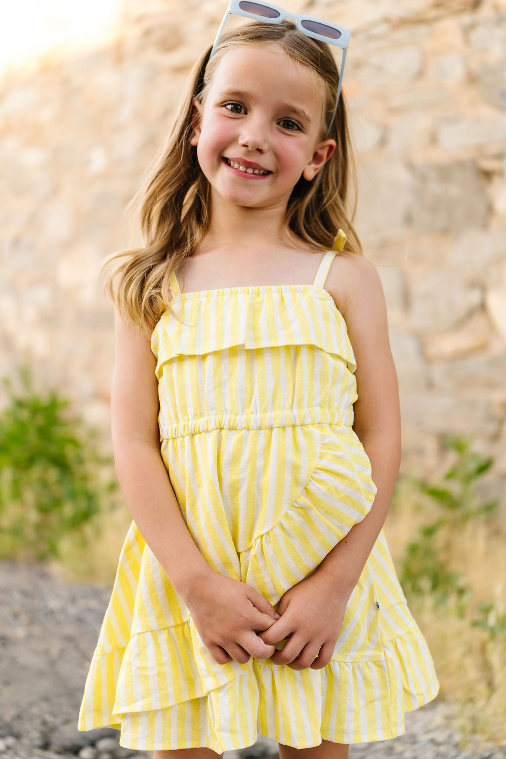 Young girl wearing a yellow striped dress with sunglasses on her head, standing against a stone wall.