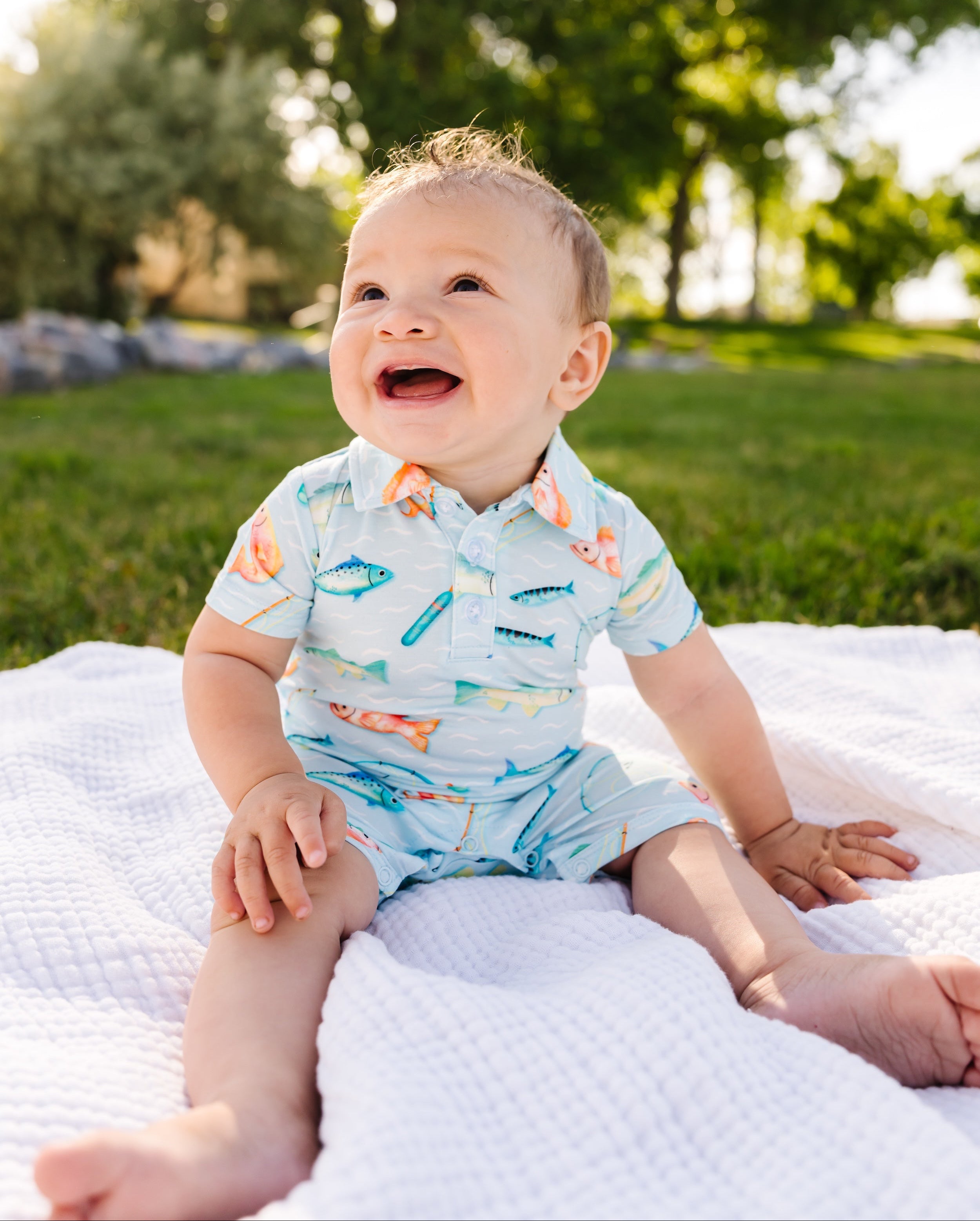 Baby sitting on a blanket outdoors with trees in the background wearing a fishing romper.