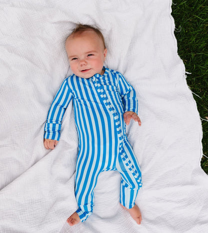 A baby lying on a white textured blanket outdoors, wearing a blue and white vertically striped one-piece pajama or romper.