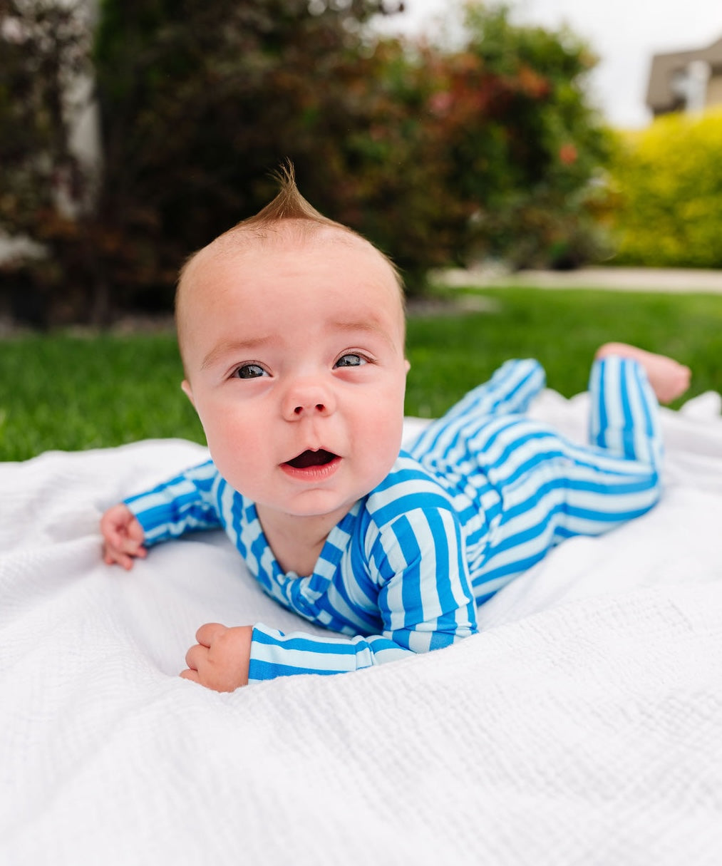 A baby lying on a white blanket outdoors, wearing a blue and white striped long-sleeve footed onesie. The background shows green grass and blurred trees.