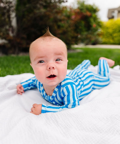A baby lying on a white blanket outdoors, wearing a blue and white striped long-sleeve footed onesie. The background shows green grass and blurred trees.