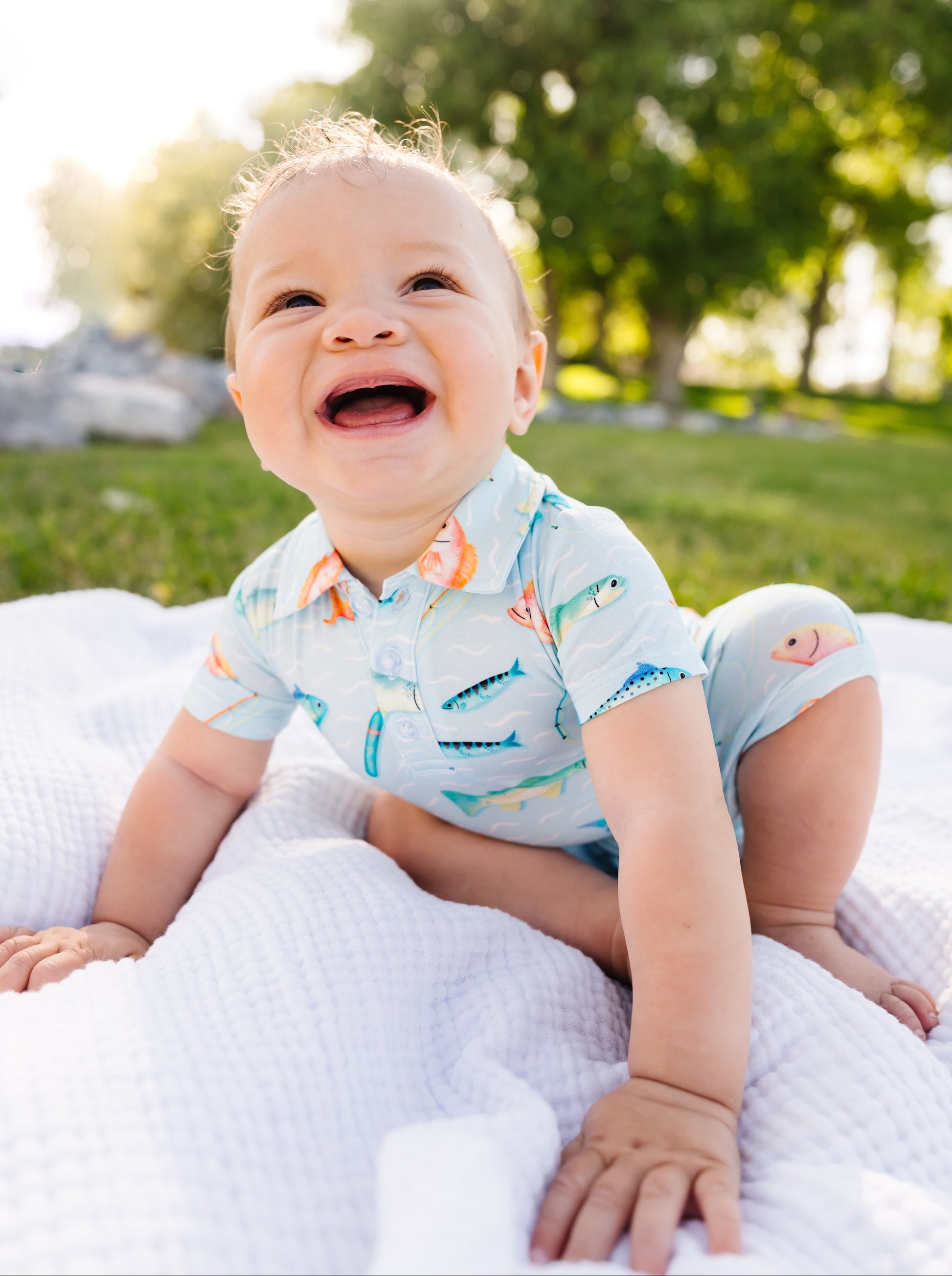 Baby sitting on a blanket in a park with trees and grass in the background