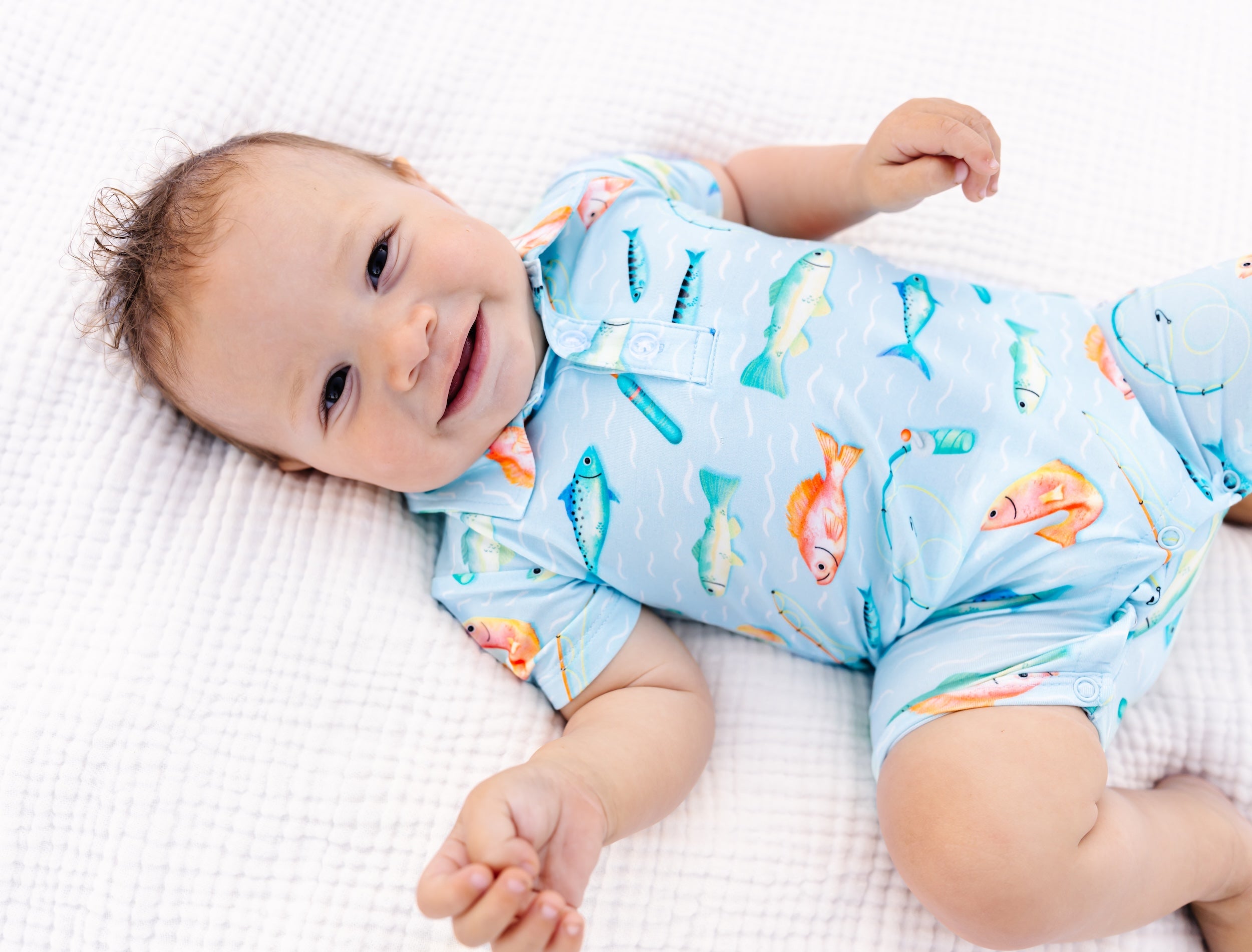 Baby wearing a colorful fish-themed polo romper lying on a white blanket