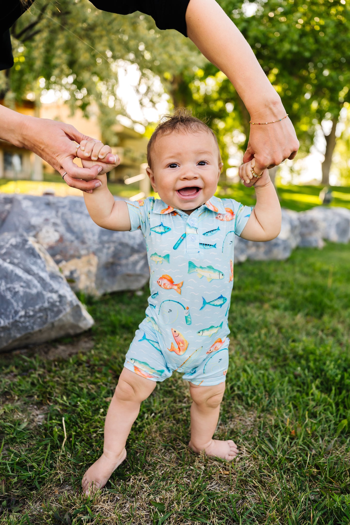 Baby in a colorful fishing themed romper standing on grass with adults holding their hands