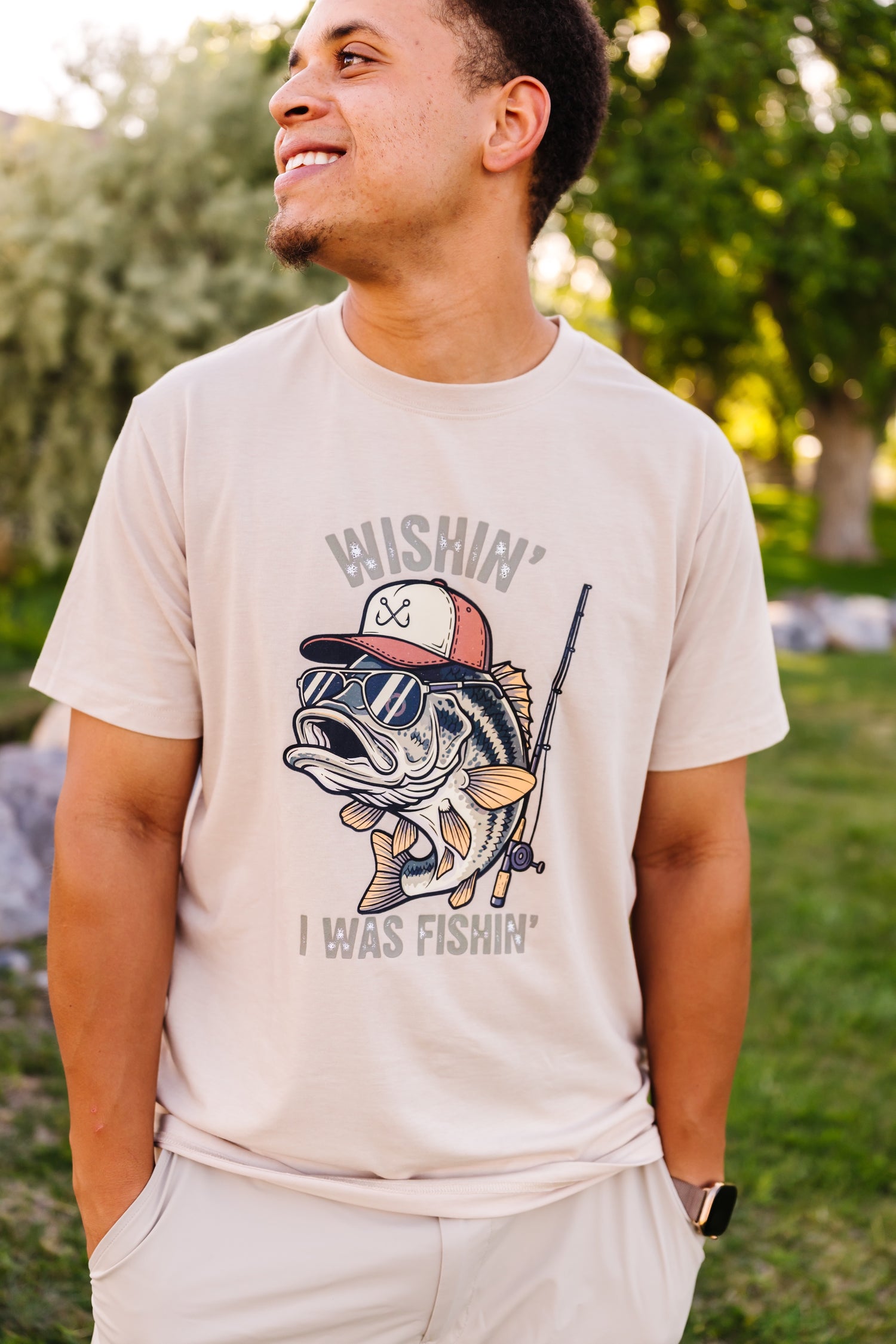 Man wearing a beige t-shirt with a graphic design of a fish holding a fishing rod outdoors.