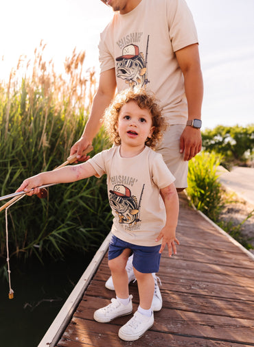 Child and adult standing on a wooden dock by a body of water, both wearing matching graphic t-shirts.